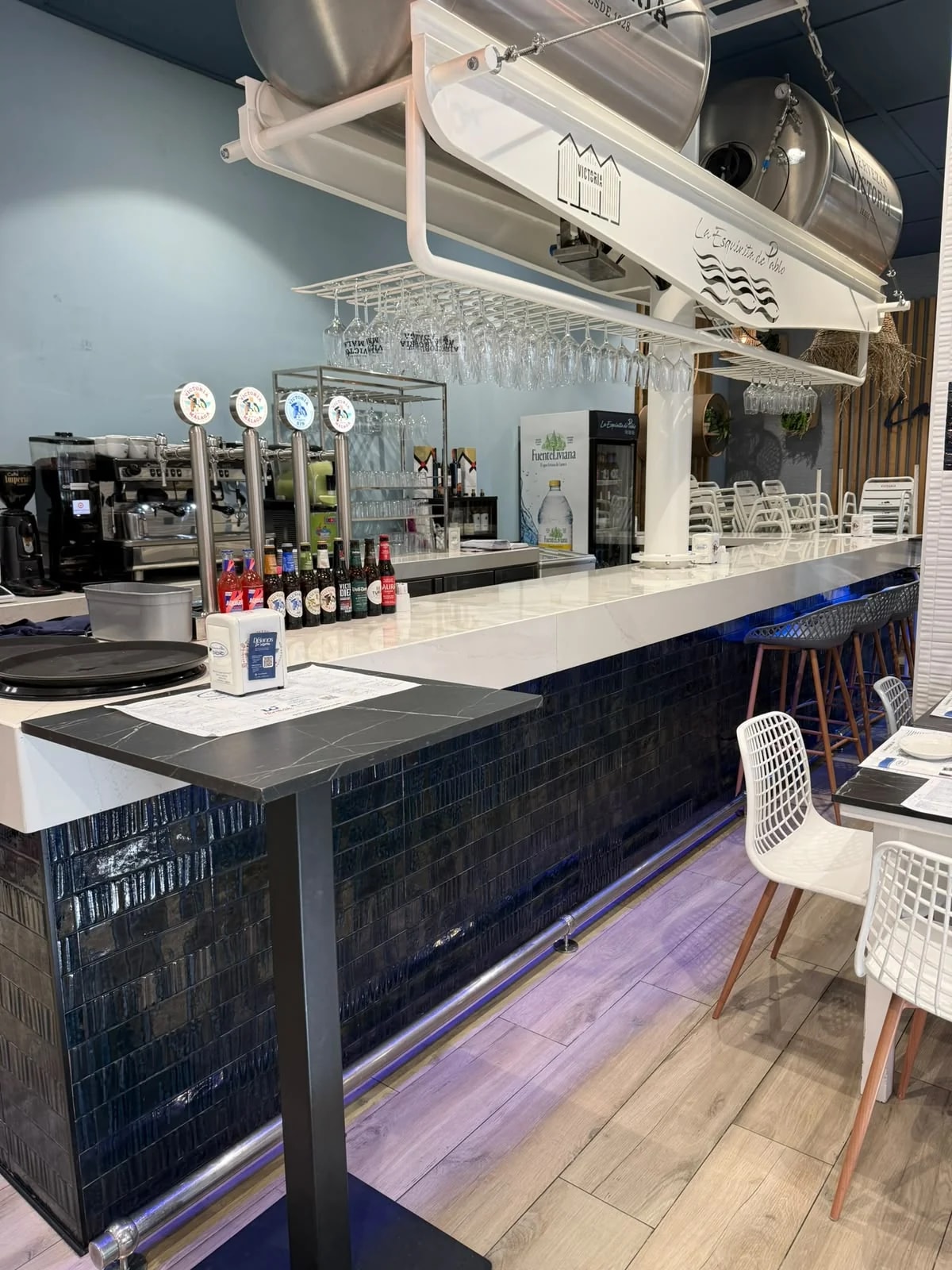 Barra del restaurante con grifos de cerveza, taburetes y estantería de copas.